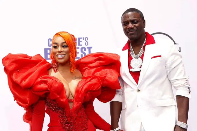 Man in white suit with red accents and gold chain necklace stands next to woman with orange hair wearing elaborate red gown with ruffled sleeves deep neckline and jewelry both smiling and posing arm-in-arm at formal event with white background and text overlay reading Greatest Night In Pop.