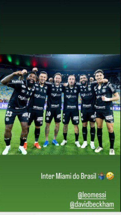 Seis jogadores de futebol sorrindo e abraçados, vestindo uniformes pretos com detalhes brancos, em um campo de grama verde sob iluminação de estádio. O primeiro à esquerda levanta o braço com o punho cerrado. Abaixo, texto Inter Miami do Brasil com emojis e menções a @leomessi e @davidbeckham