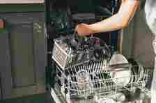 A person loads a dishwasher by placing a cutlery basket inside. The open dishwasher has various dishes and glasses