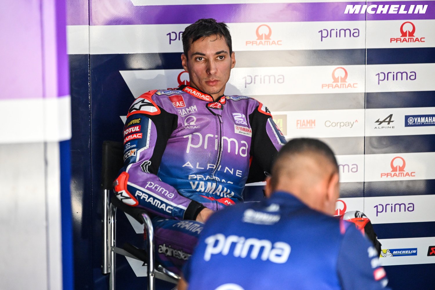 Rider in purple Prima Pramac Yamaha MotoGP leathers sits in the garage beside a team member in blue, with sponsor boards behind.