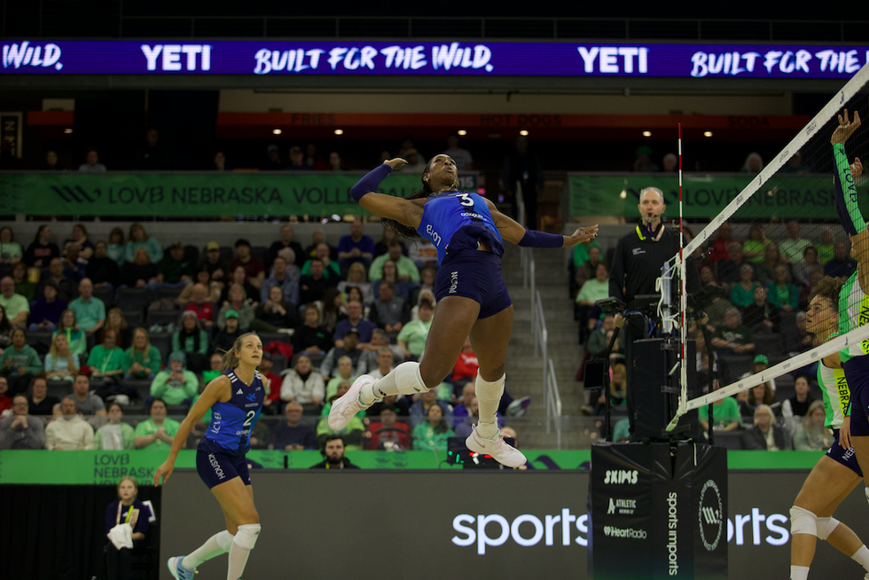 A volleyball player leaps to serve while two teammates and spectators watch in the background.