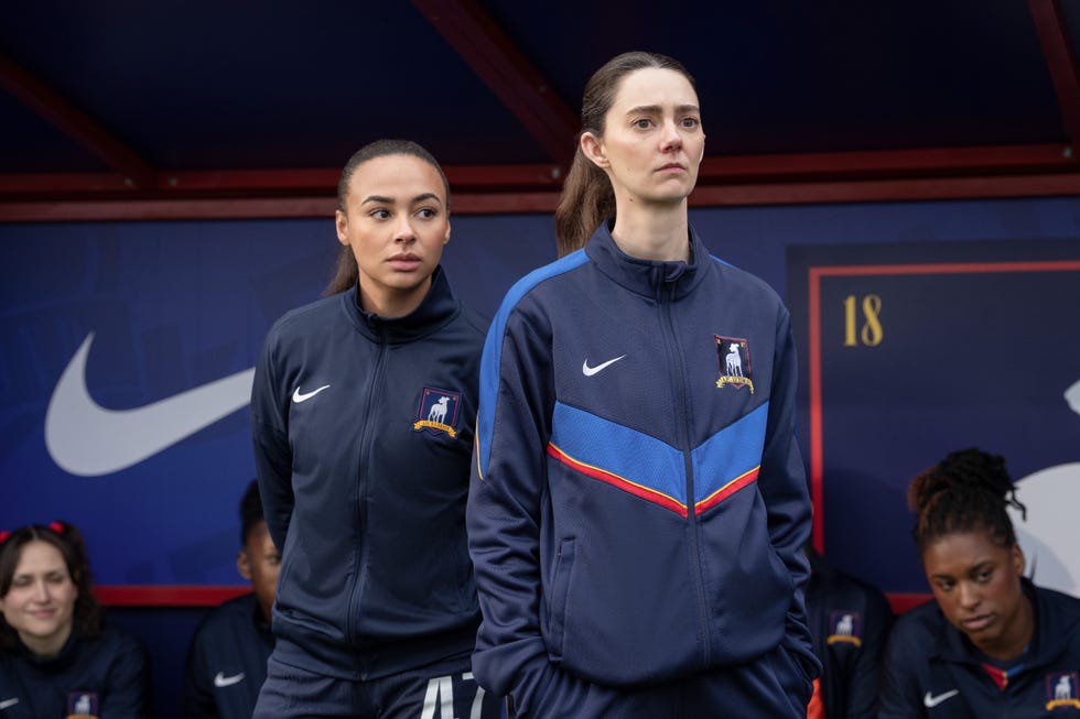 Two athletes in sports jackets stand on the sideline, showing focus and determination during a team event.