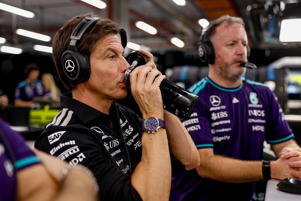 Man in black Mercedes team shirt and headphones drinking from a bottle in the pit garage, Formula 1 team crew nearby
