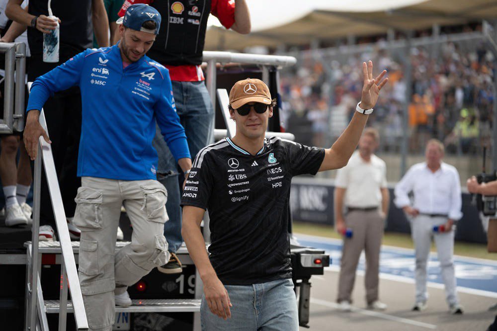 Man in Mercedes black shirt and cap waving on a paddock walkway, with Alpine-clad staff nearby at an F1 event