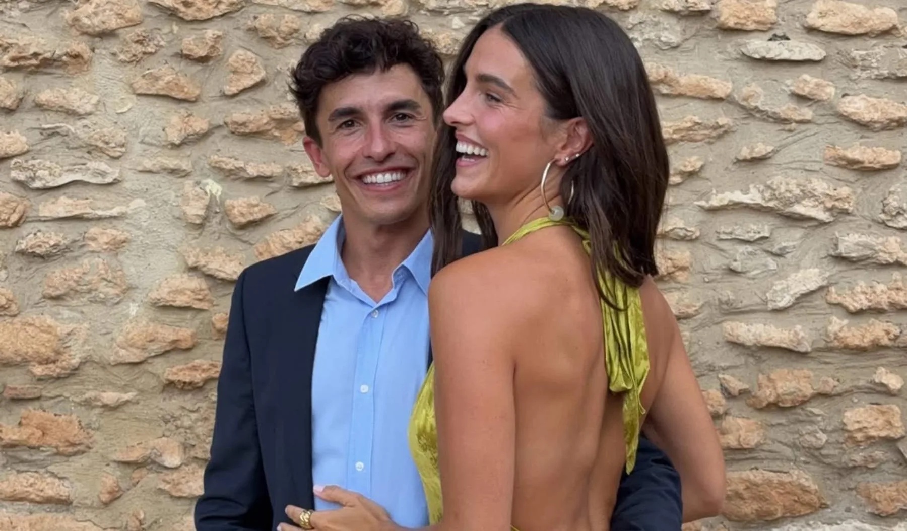 Two smiling people embrace in front of a rustic stone wall; a man in a navy jacket and blue shirt and a woman in a yellow halter top laugh together.
