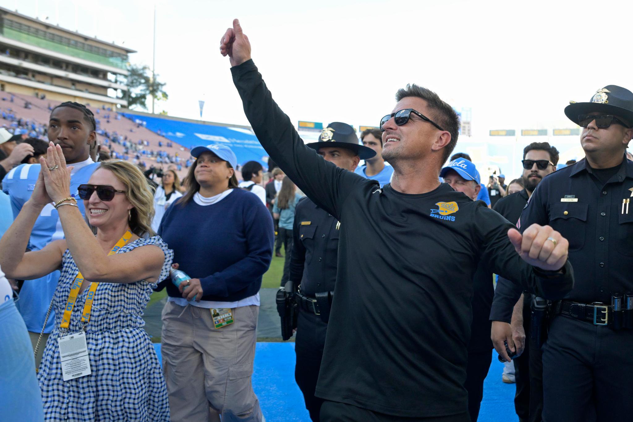 A man in a black long-sleeved UCLA shirt raises his arm in celebration, surrounded by fans and security personnel at a stadium.