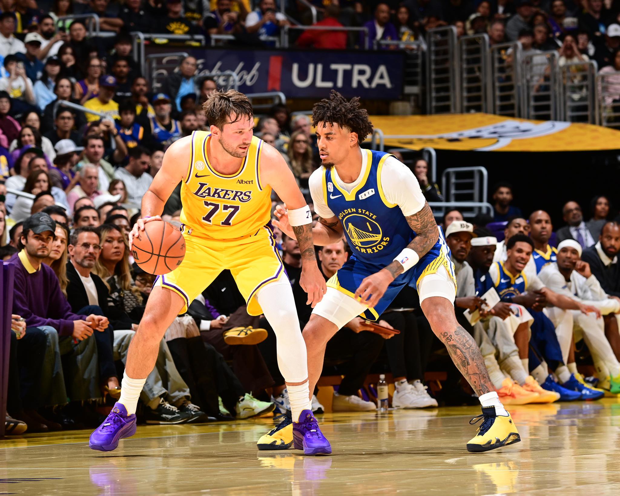 Luka Doncic in a Lakers uniform dribbles the basketball while being guarded by a Golden State Warriors player.