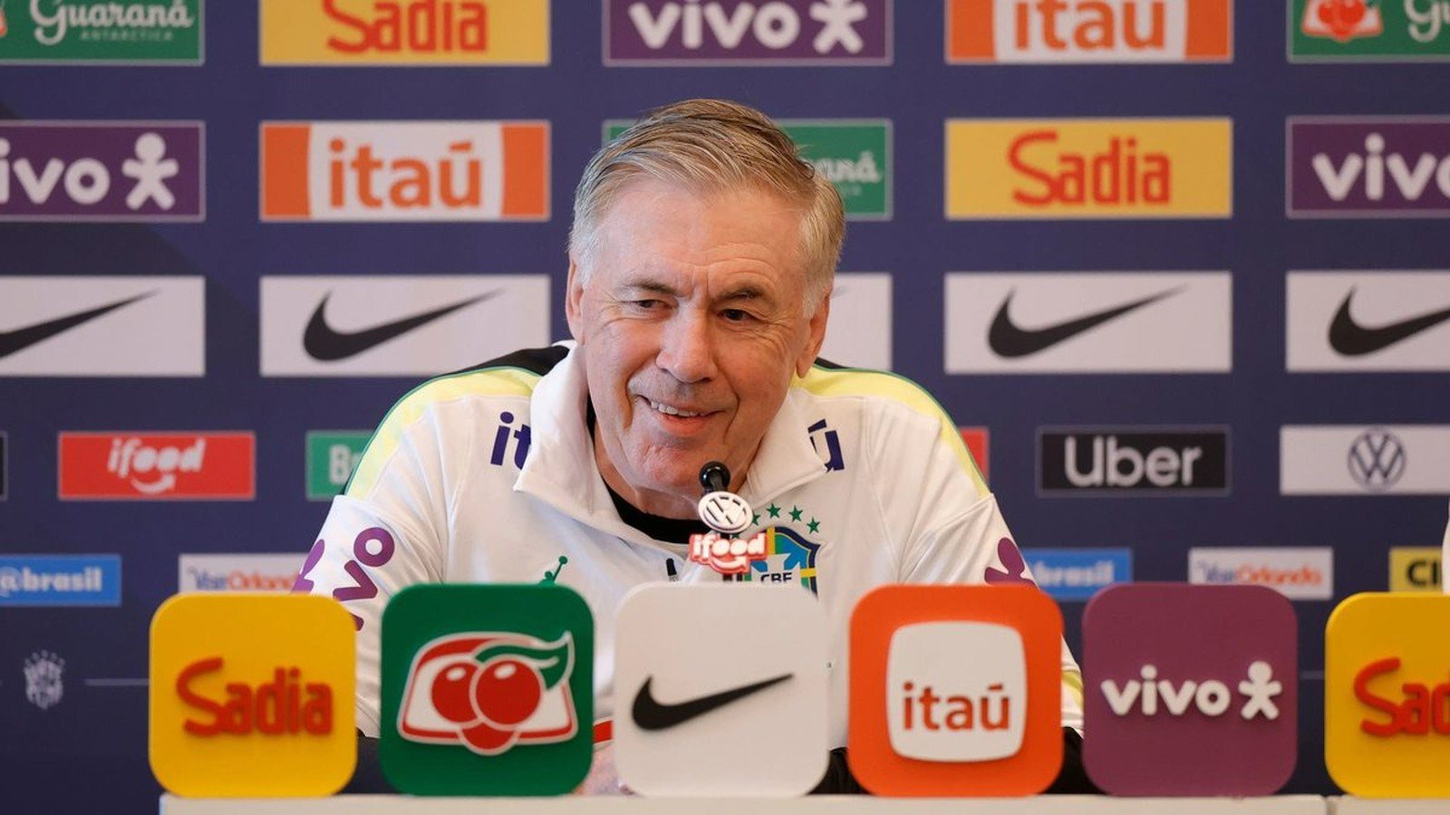 Carlo Ancelotti sorrindo em coletiva de imprensa com vários logotipos de patrocinadores