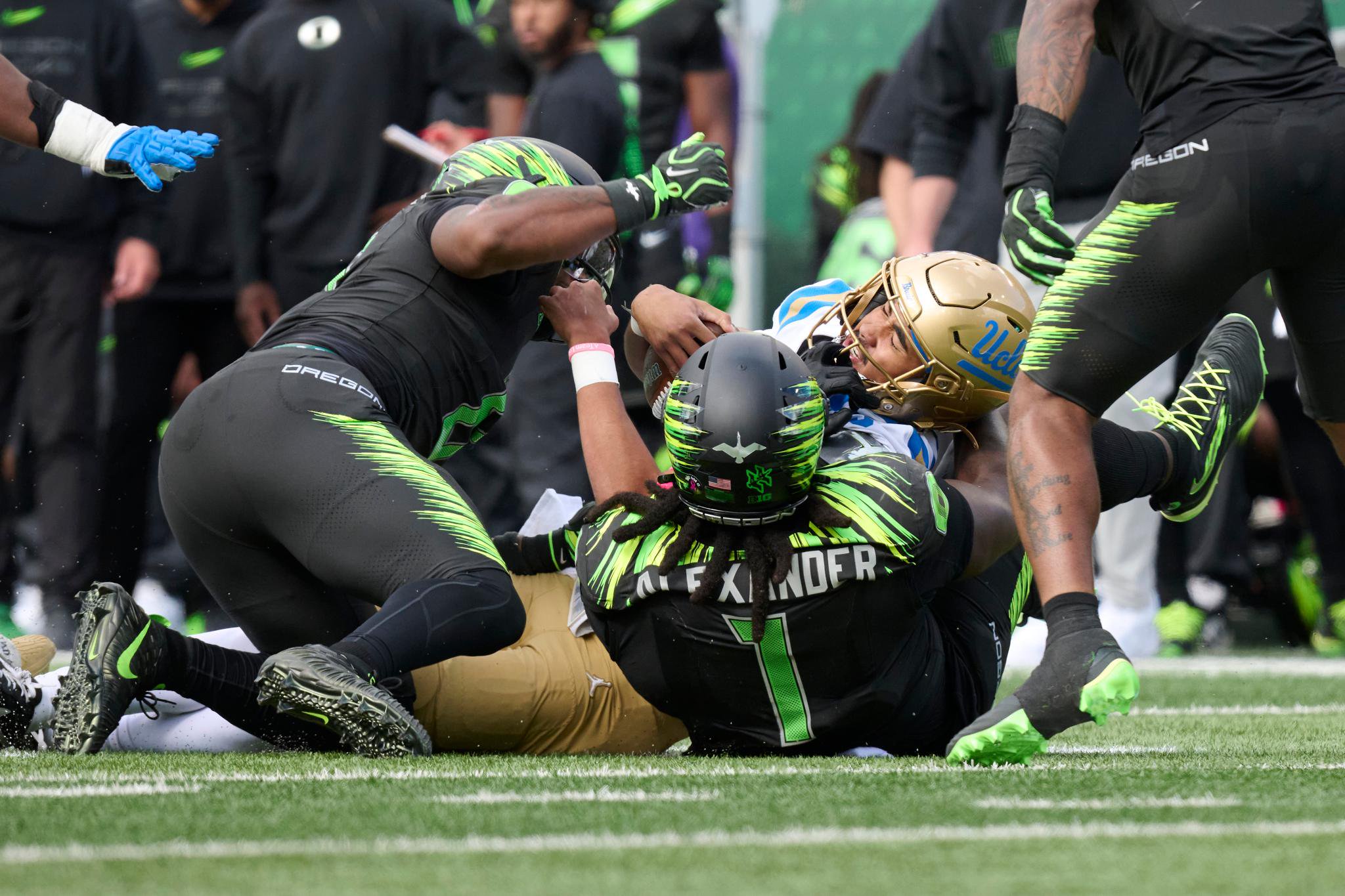 A UCLA football player in a gold and blue uniform is tackled by multiple Oregon players wearing black and green uniforms on the field.