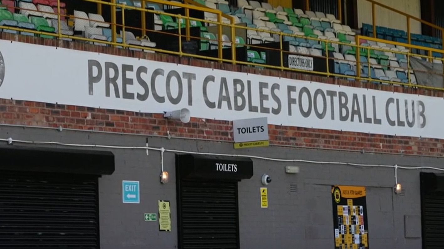 A stand in a football ground featuring a number of plastic seats over a brick structure. A white sign stretches across brickwork below the seating area which reads: 'Prescot Cables Football Club' in black lettering. 