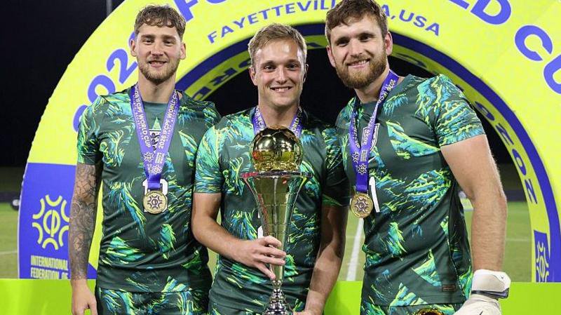 Reagan McMillan, Freddie Lamb and Giles Moore of England, all dressed in green goalkeeping kits, pose for a photos with the trophy following the team's victory