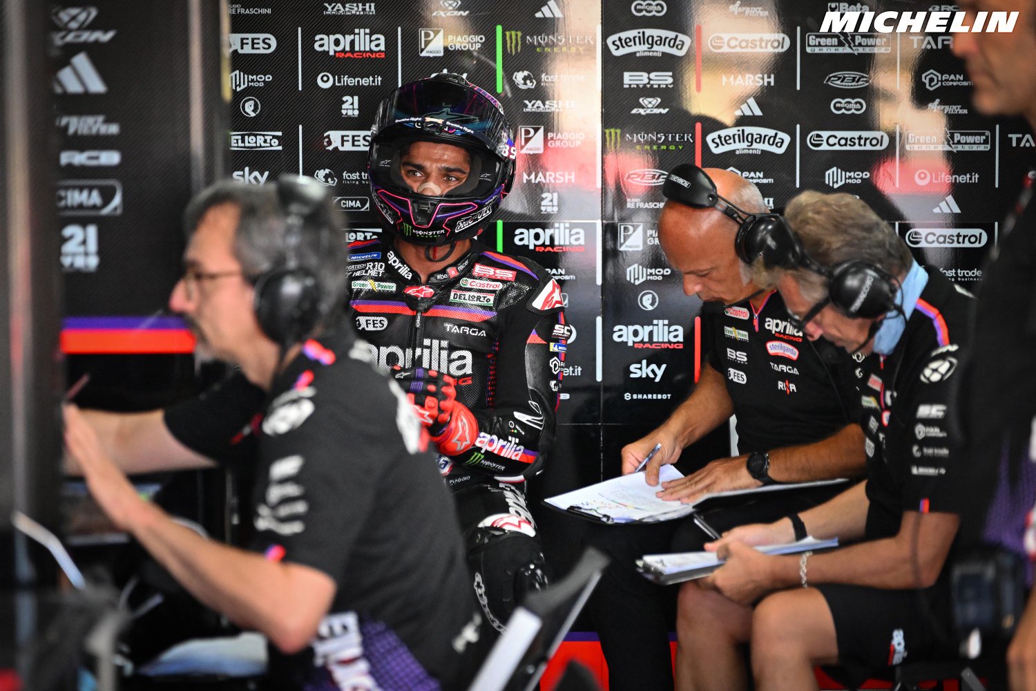 Jorge Martin in Aprilia racing gear and helmet in the pit garage, with team crew members wearing headsets during MotoGP.