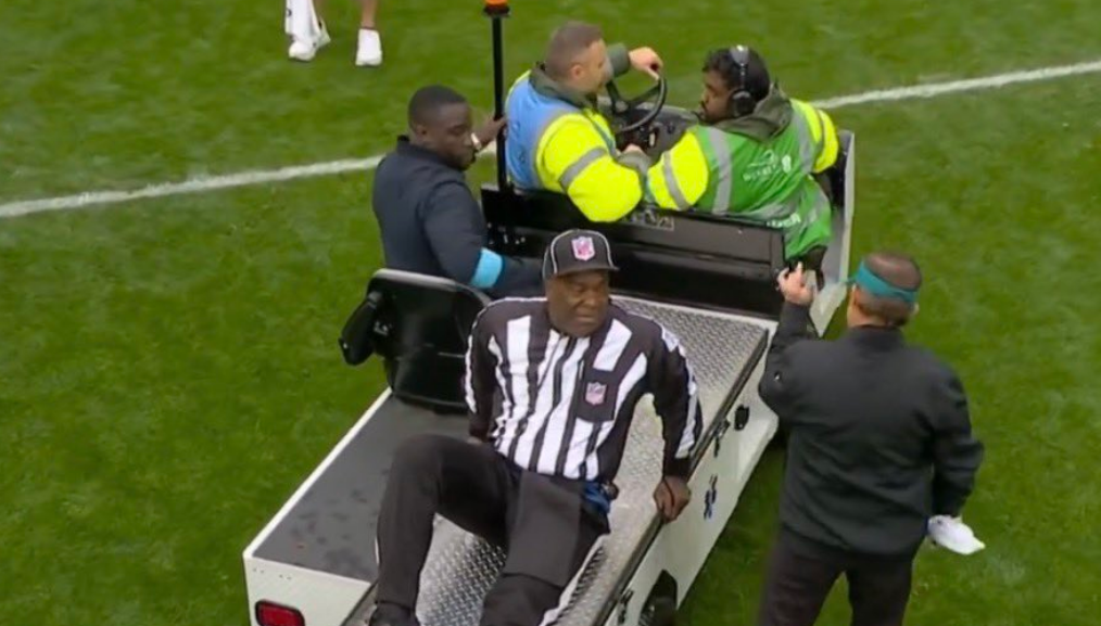 An NFL referee sits on the back of a medical cart while another person stands nearby, pointing.