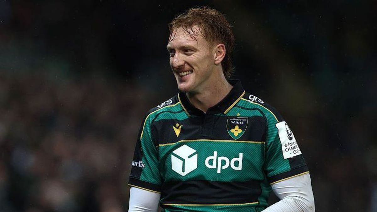 George Hendy standing on the pitch during a Champions Cup match for Northampton. He is wearing a green and black home kit with a white long-sleeved top underneath