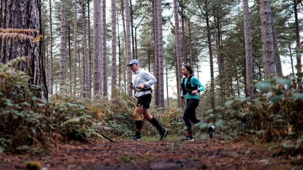 A man and a woman running through a wooded area. The picture is taken from ground level. The woman is wearing a blue top with black running vest and black leggings, while the man wears black shorts and a white windbreaker and light blue cap.