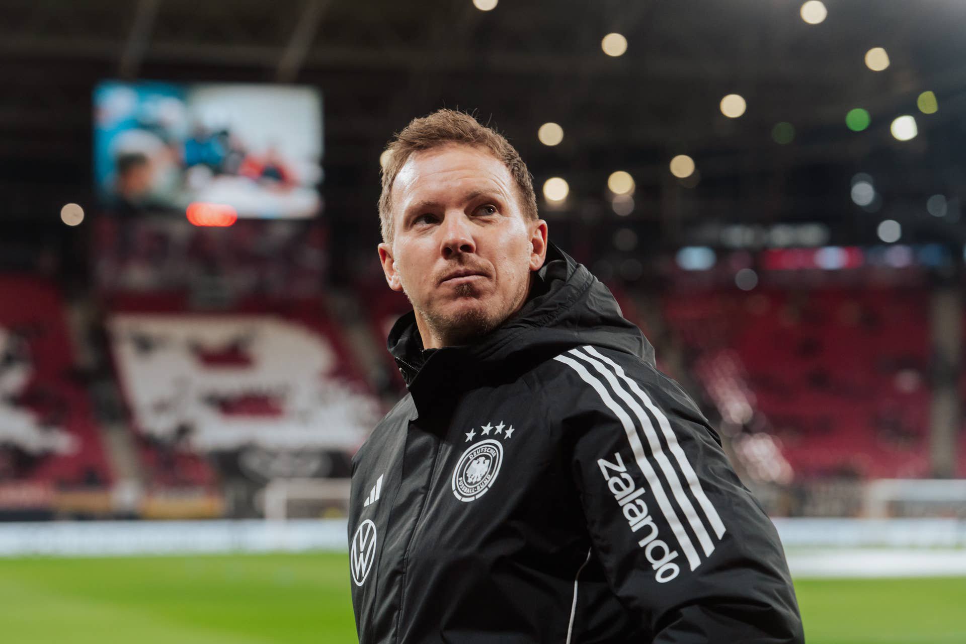 Ein Fußballtrainer im Deutschland-Trainingsanzug blickt konzentriert im Stadion nach oben.