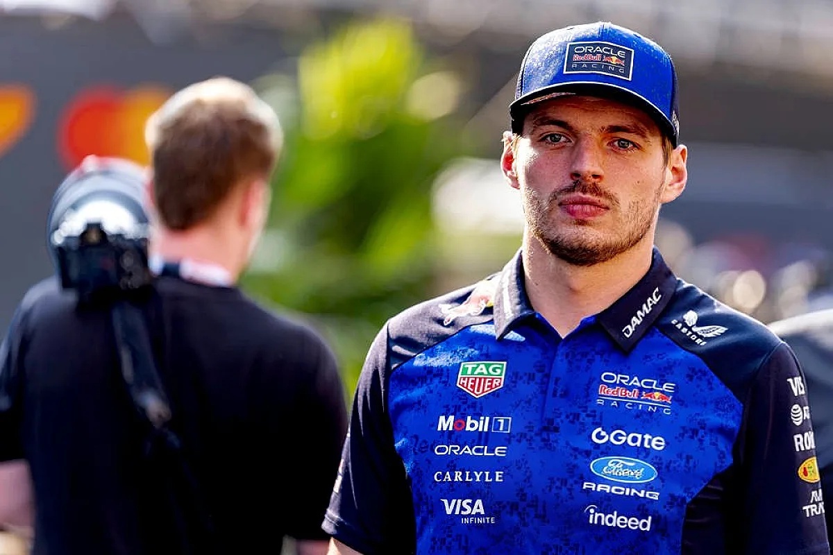A Red Bull Racing driver in a blue team shirt with Oracle, Tag Heuer and Visa logos stands in a paddock, looking ahead while a crew member stands nearby.