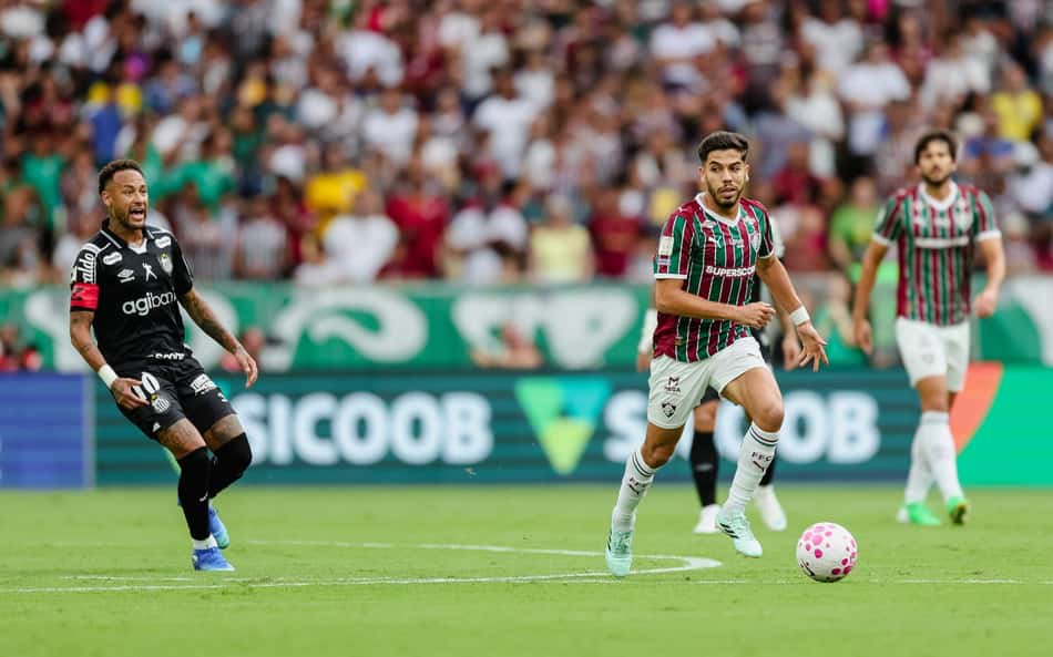 Jogador do Fluminense com a bola em ataque veloz, cercado por adversário com uniforme preto, estádio lotado ao fundo.