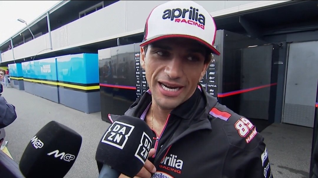 Rider in an Aprilia Racing cap and jacket speaking to DAZN and MotoGP microphones in a paddock, Phillip Island