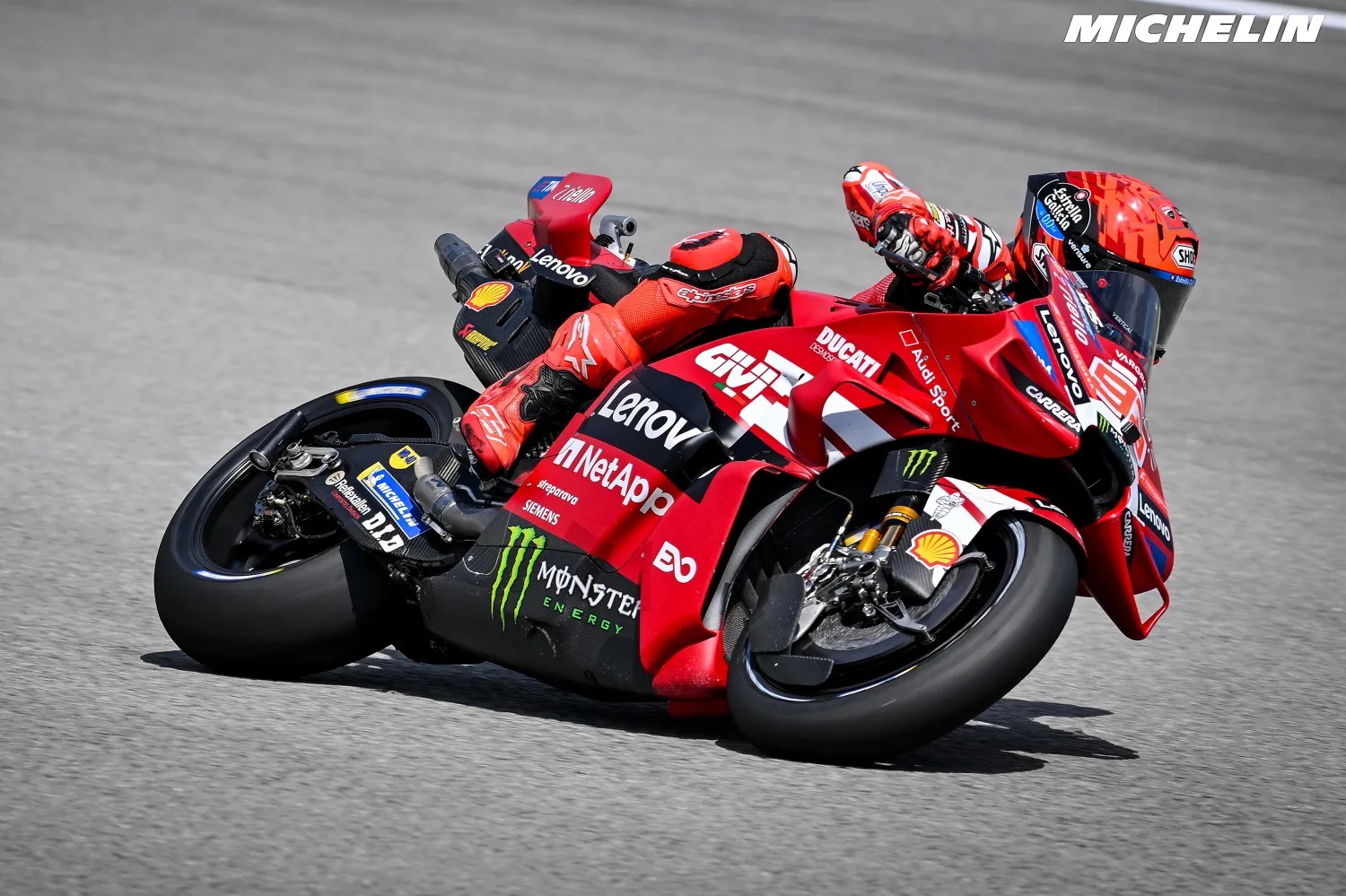 A rider in red Ducati leathers and helmet leaning deep into a corner on a red Ducati MotoGP bike, with Lenovo and Monster livery visible, on the track.