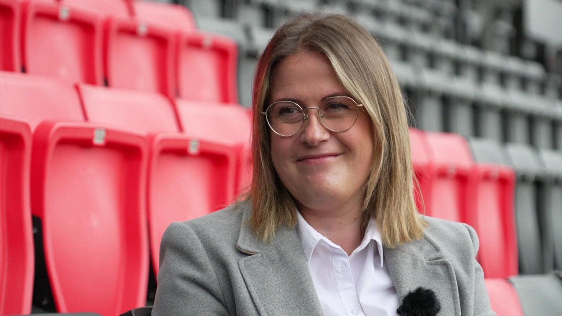 Councillor Millie Earl sits in the black and red stands of Vitality Stadium for an interview. She has shoulder length, straight blonde hair and round metal-rimmed glasses. She is wearing a pale grey suit jacket and and white shirt with an open collar.