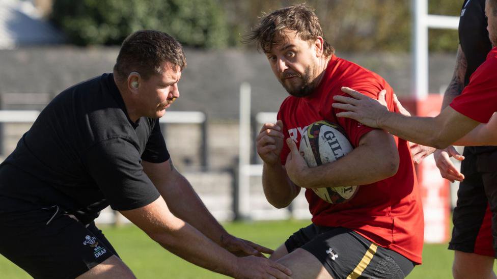 Ryan Shallish (left) is wearing all black t-shirt and shorts and wears white hearing aids tackles Joe Yeates (centre) who is wearing a red sports t-shirt, black rugby shorts and is hugging the rugby ball during a Wales Deaf Rugby training drill, while Andrew Healy (right back) wearing all black t-shirt and shorts and Anthony Leach (right front) wearing red t-shirt and green shorts provide support