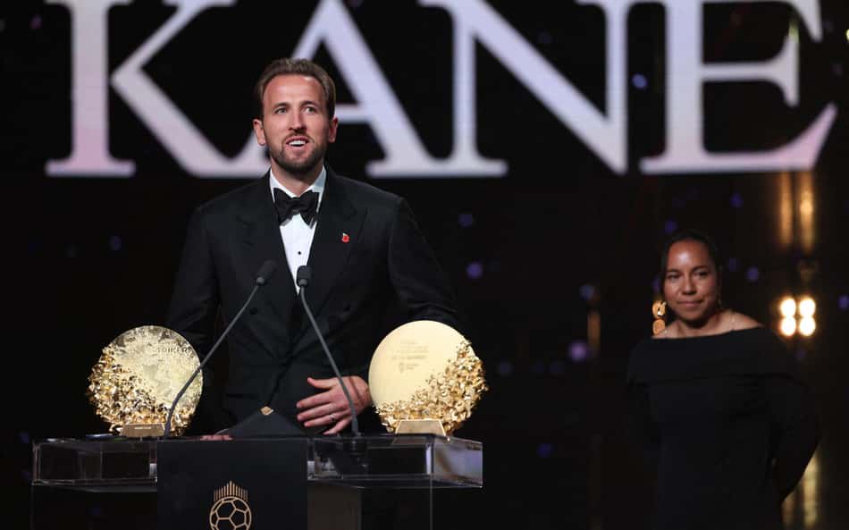 Homem em smoking no palco da premiação Kane, com dois troféus dourados, mulher de preto ao fundo.