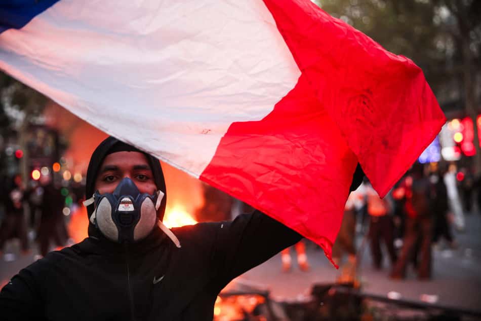 Manifestante usando máscara de gás segura uma bandeira francesa em frente a um incêndio no Boulevard des Capucines, durante manifestação contra o alto custo de vida em Paris, na França, em 17 de outubro de 2026.