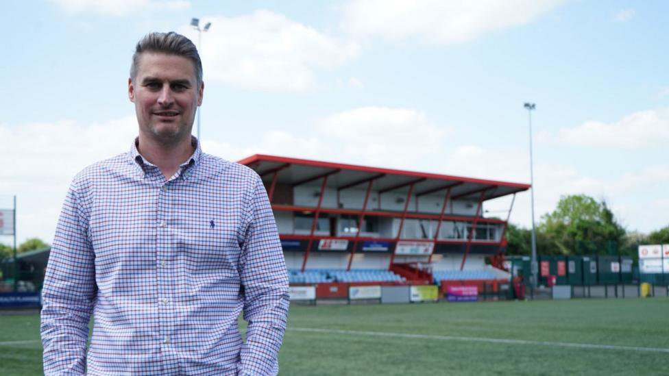 David Faulkner stands inside a football ground, with the stands behind him. He is in a red and blue checked long sleeve shirt.