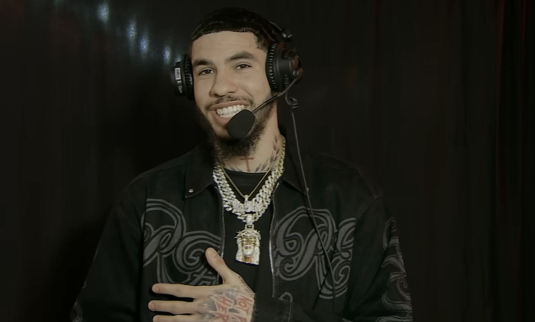 Person speaking with headphones and mic, wearing a jacket with patterned designs and multiple necklaces
