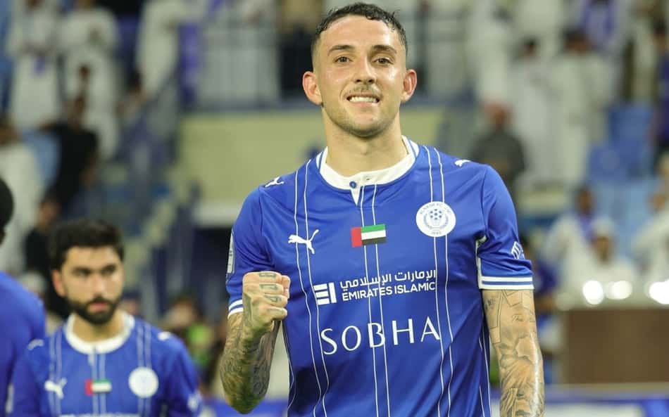 Jogador de futebol em camisa azul com listras brancas, bandeira dos Emirados Árabes Unidos no peito, em estádio.