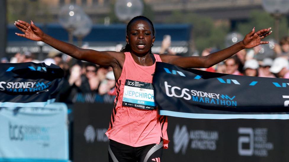 Peres Jepchirchir in a pink top with arms outstretched, breaking a ribbon as she crosses the finish line at the Sydney marathon