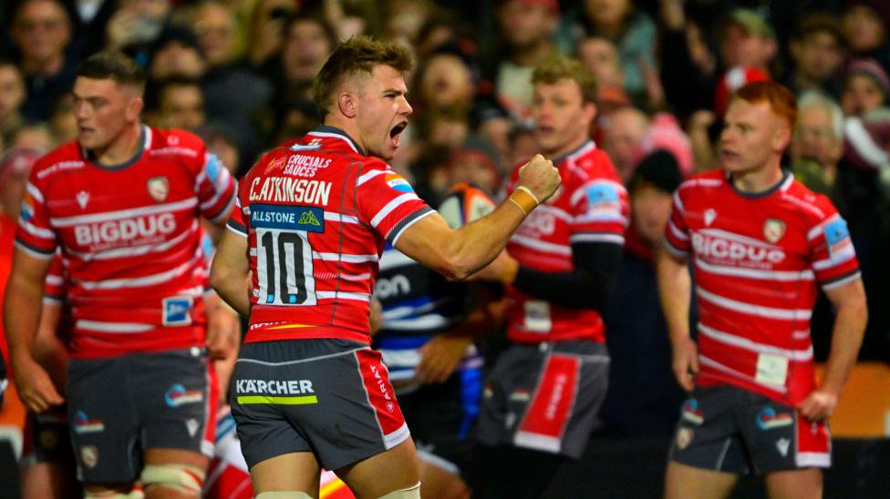 Gloucester players celebrate stopping Bath scoring a try. The Gloucester players are wearing cherry red shirts with thin white horizontal stripes. The player in the middle of the picture is shouting and punching the air in celebration. 