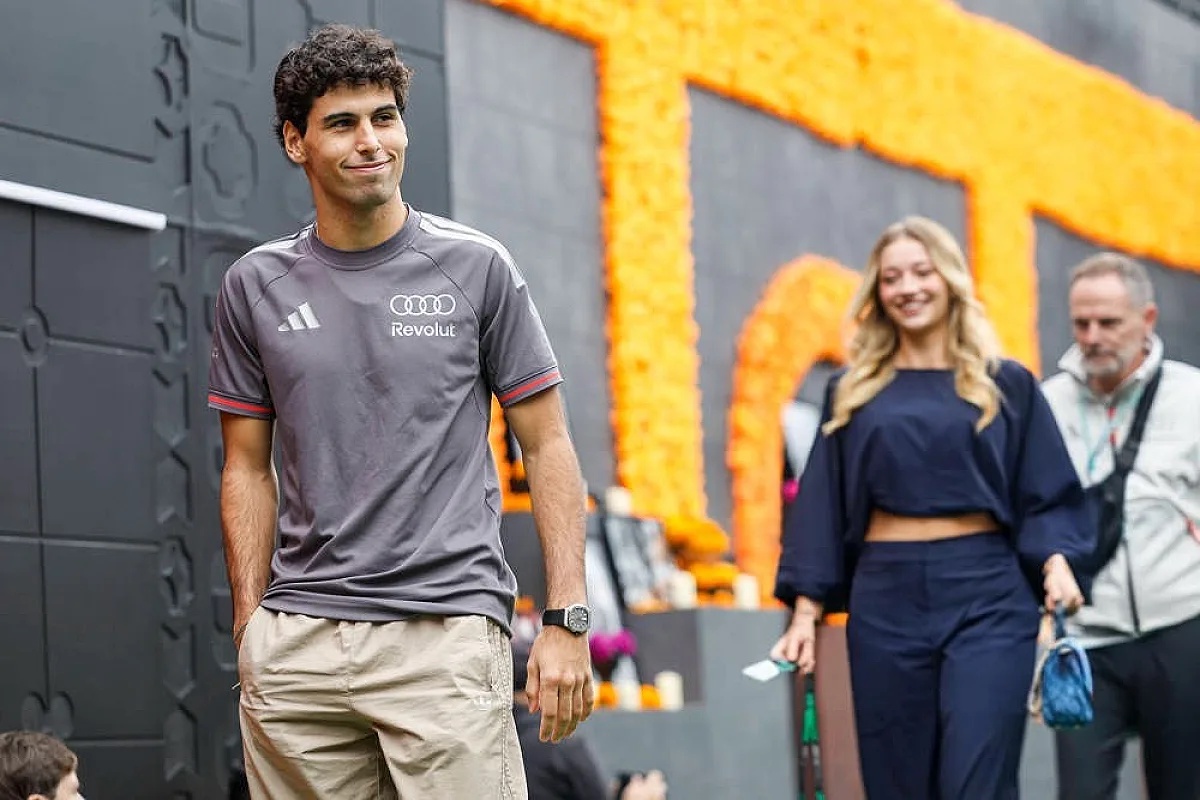 A young man in a grey Audi Revolut-branded adidas shirt and beige shorts smiles while walking past an orange floral wall in the paddock.