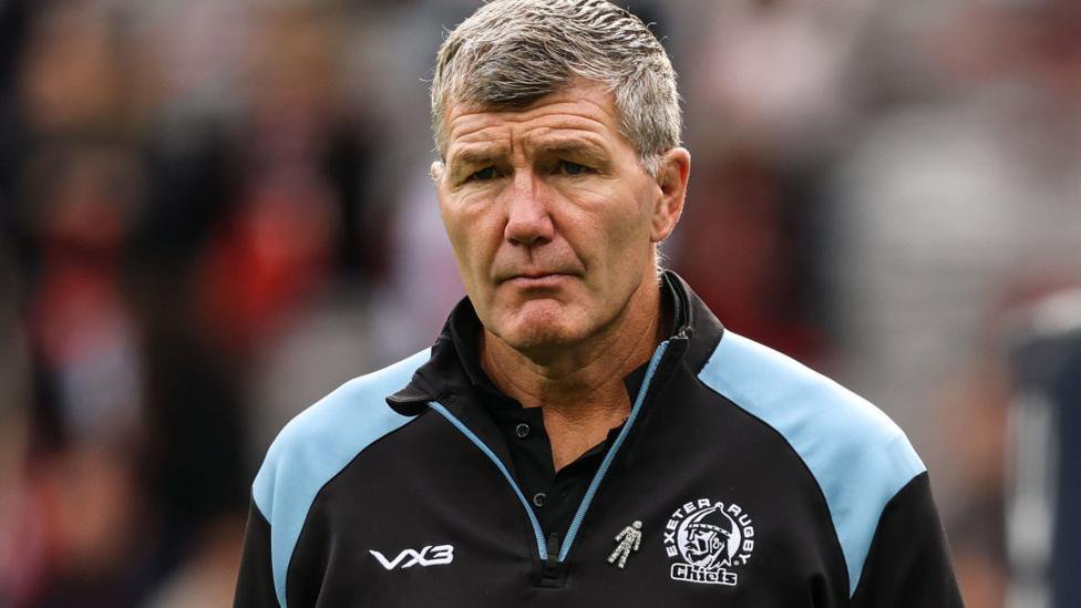 Rob Baxter looks straight ahead while standing on the pitch of a Prem match. He is wearing a black and blue Exeter Chiefs branded top