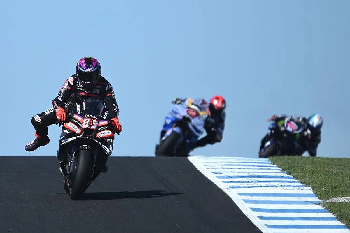 Aprilia MotoGP rider in black and red leathers, number 89, lifting a wheel on the track with rivals behind him