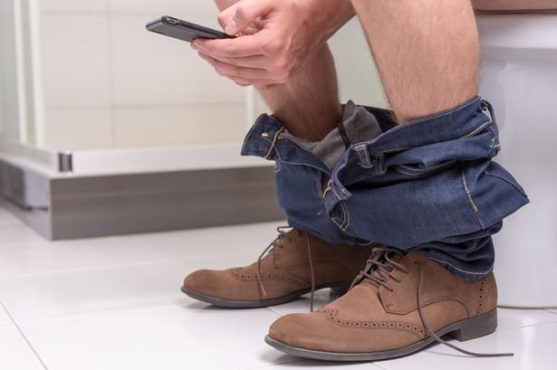 Shot of a man's boots and pulled down trousers as he sits on the toilet on his phone