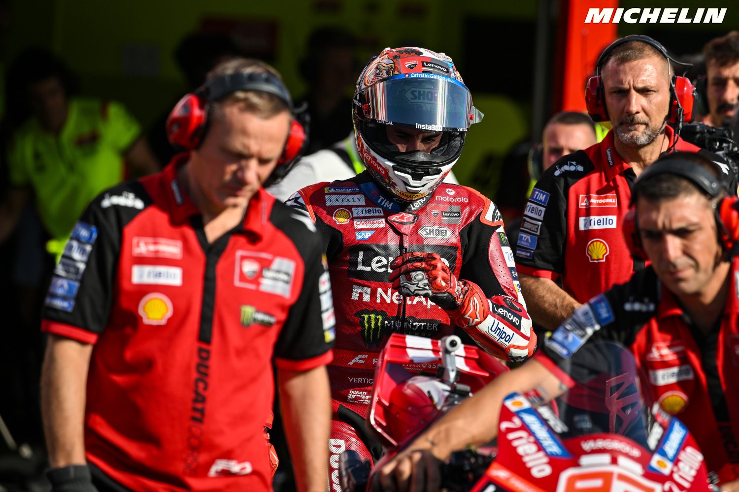 A MotoGP rider in a red Ducati leathers and helmet stands in the pit lane beside a red Ducati motorcycle, with team crew in red shirts around him