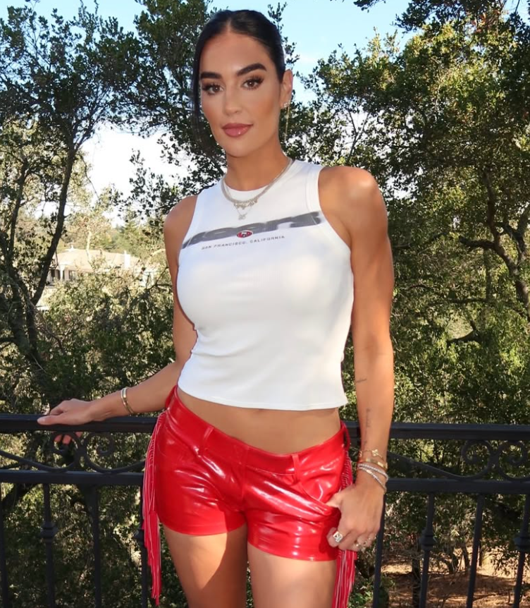A woman standing outdoors on a balcony wearing a white tank top and red shorts.