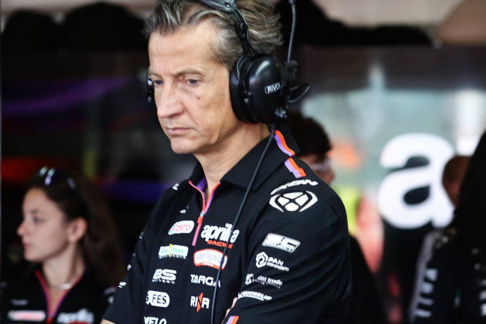 A man in a black Aprilia Racing team shirt with headphones in the pit garage, looking down intently, near a team member