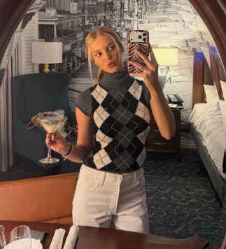 A blonde woman wearing an argyle sweater vest and white pants takes a mirror selfie while holding a martini glass.