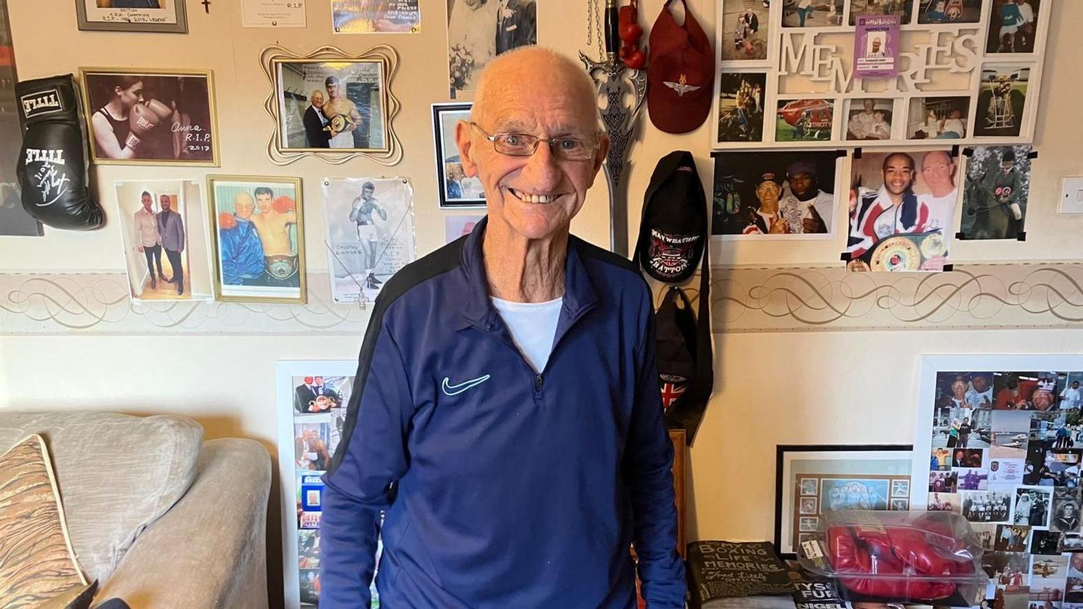 A man in his 90s standing next to a wall full of pictures and artwork from throughout his boxing career.