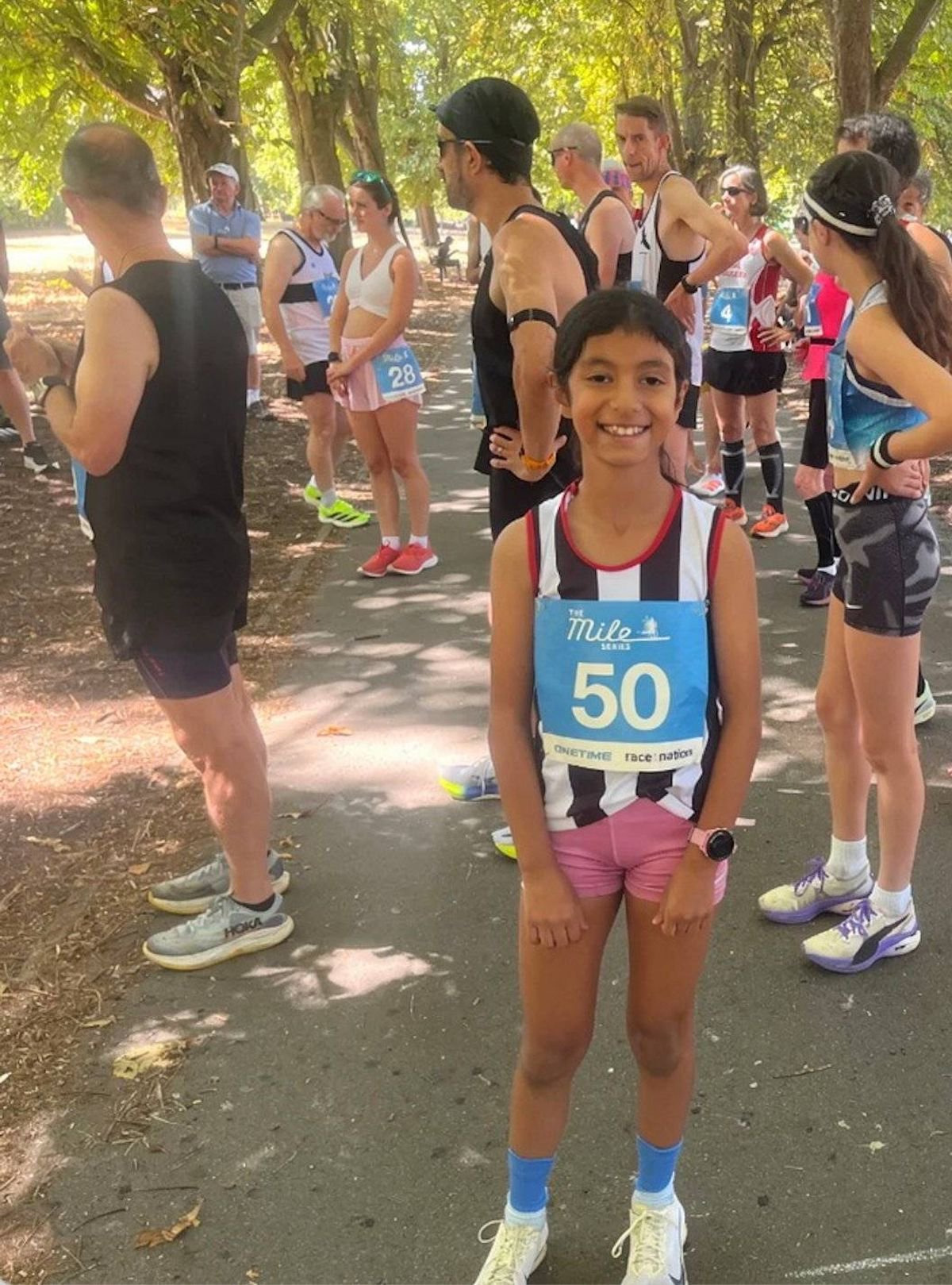 A young individual, wearing a race bib numbered 50, stands in the midst of a group of runners on a pathway surrounded by trees. The individuals are dressed in various athletic gear, including bibs, sneakers, and caps, suggesting the setting of a running event.