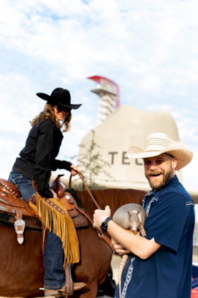 Cowgirl next to a man holding an armadillo in front of the largest cowboy hat