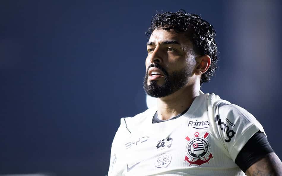 Jogador do Corinthians em uniforme branco, com o escudo do clube, em campo sob luzes, expressão concentrada.