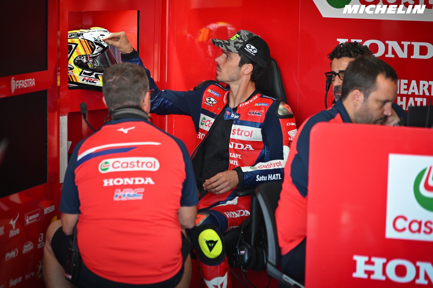 Rider in Honda HRC red-and-navy leathers seated in the garage, reaching up toward a helmet, with Castrol and Honda crew nearby