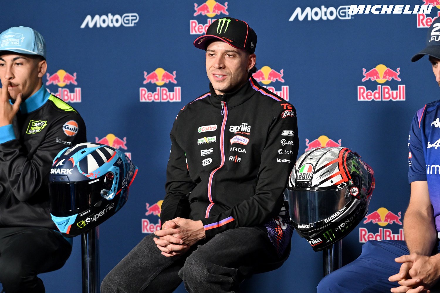 Three riders at a MotoGP press conference in front of Red Bull and Michelin banners; the centre rider wears an Aprilia Racing jacket and Monster cap, with he…