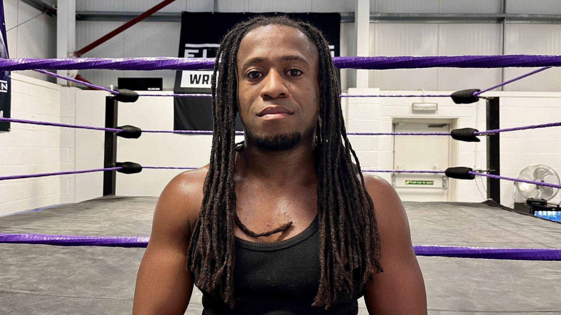 A 28-year-old man with long dreadlocks looks into the camera. He has a stern expression and is wearing a black vest. Behind him is a wrestling ring.