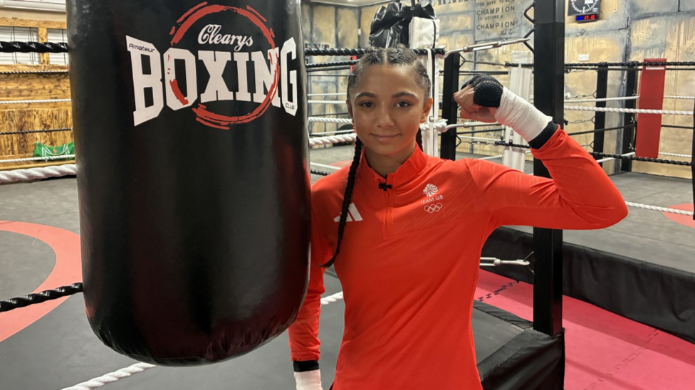 A girl wearing a orange top stands outside a boxing ring, with her arm up in a fighting stance.