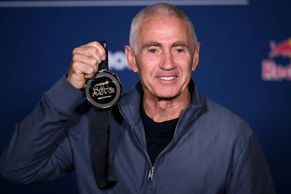 Mick Doohan holding a MotoGP Hall of Fame medal at Phillip Island, smiling in a blue zip jacket against a dark blue backdrop.